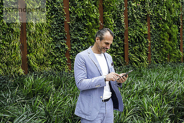 Mature businessman text messaging on mobile phone standing in front of hedge