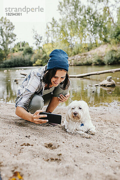 Lächelnde junge Frau macht ein Selfie mit Hund am Seeufer im Park