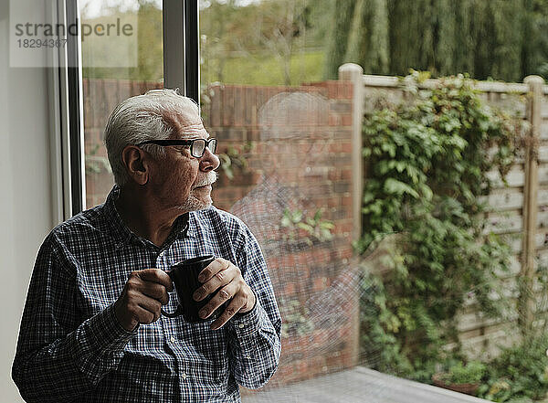 Nachdenklicher älterer Mann mit einer Tasse Kaffee  der zu Hause aus dem Fenster schaut