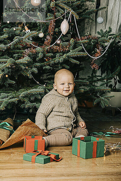 Lächelnder kleiner Junge mit Geschenkboxen sitzt vor dem Weihnachtsbaum