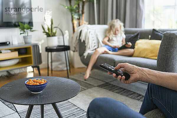 Hand of man holding remote control with daughter in background on sofa
