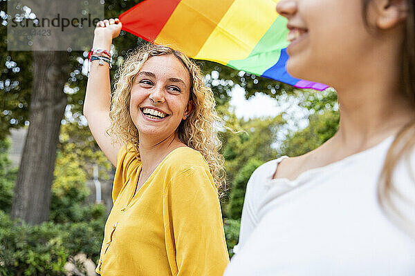 Glückliche junge Frau mit Regenbogenfahne und Blick auf Freundin im Park