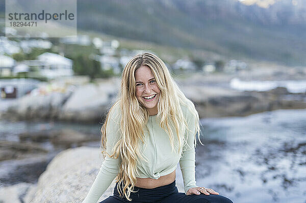 Glückliche junge Frau mit blonden Haaren sitzt auf einem Felsen