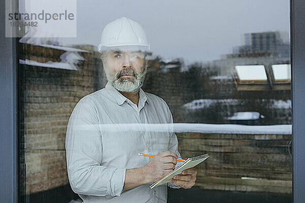 Nachdenklicher Architekt mit Schutzhelm steht vor einer Molkerei  die durch Glas gesehen wird