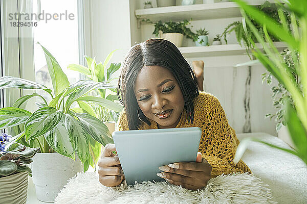 Lächelnde Frau mit Tablet-PC  die zu Hause am Fenster liegt