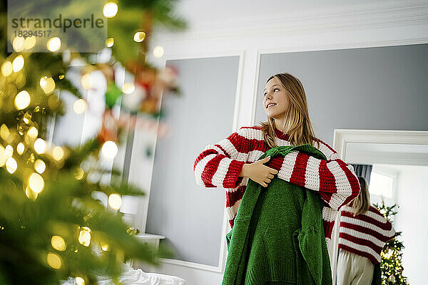 Smiling girl holding green sweater with Christmas tree in foreground