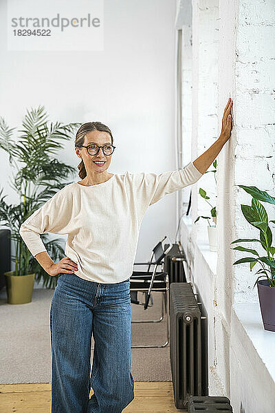 Nachdenkliche reife Geschäftsfrau mit der Hand auf der Hüfte  die an der Wand im Büro steht