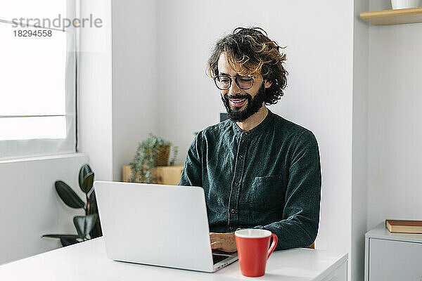 Lächelnder bärtiger Geschäftsmann benutzt Laptop am Schreibtisch und arbeitet im Büro