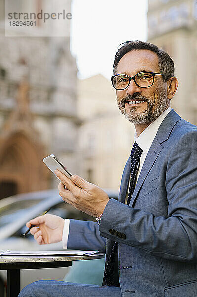Smiling mature businessman with smart phone sitting at cafe