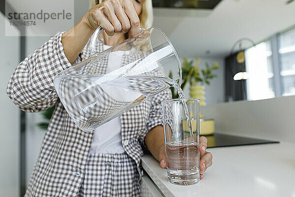 Durstige Frau gießt zu Hause Wasser in Glas durch Glas