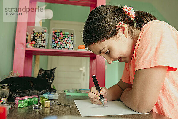 Lächelndes Mädchen  das auf Papier zeichnet  während die Katze am Tisch sitzt