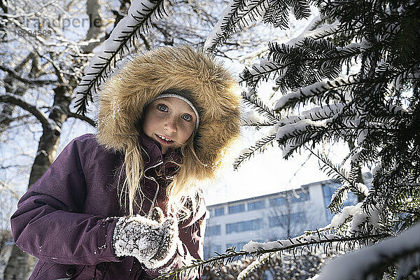 Lächelndes Mädchen mit Pelzmütze  das unter einem Tannenbaum steht