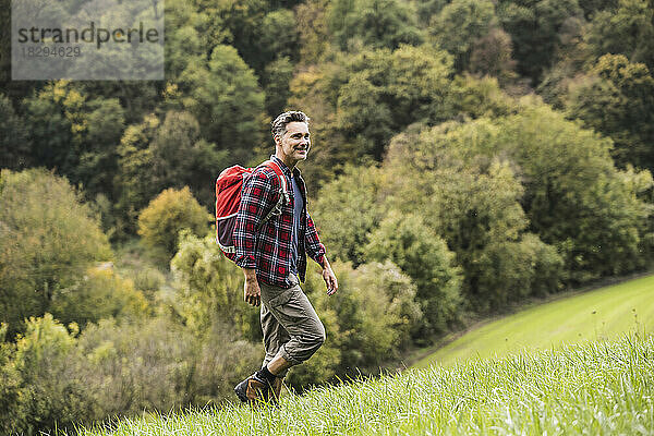 Lächelnder reifer Mann mit Rucksack  der auf Gras vor Bäumen läuft