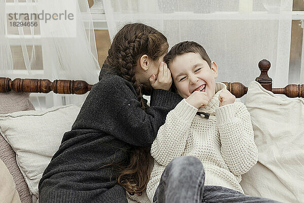 Sister whispering in brother's ear on sofa at home