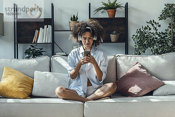 Junge Frau surft mit Mobiltelefon im Internet und sitzt zu Hause auf dem Sofa