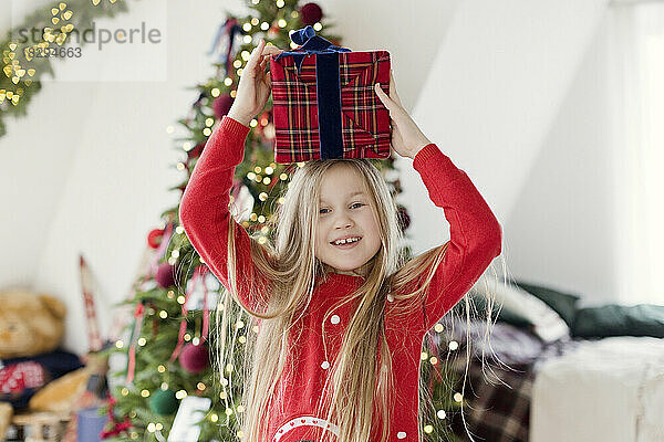 Fröhliches blondes Mädchen hält zu Hause ein Weihnachtsgeschenk auf dem Kopf