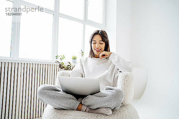 Glückliche Frau sitzt mit gekreuzten Beinen zu Hause und benutzt Laptop