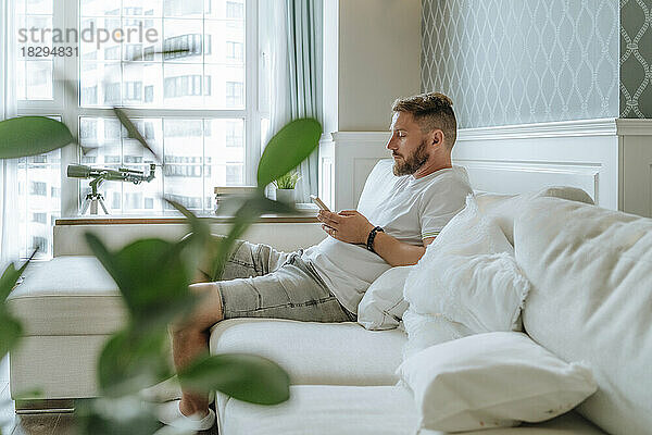 Junger Mann mit Bart benutzt Smartphone auf Sofa zu Hause