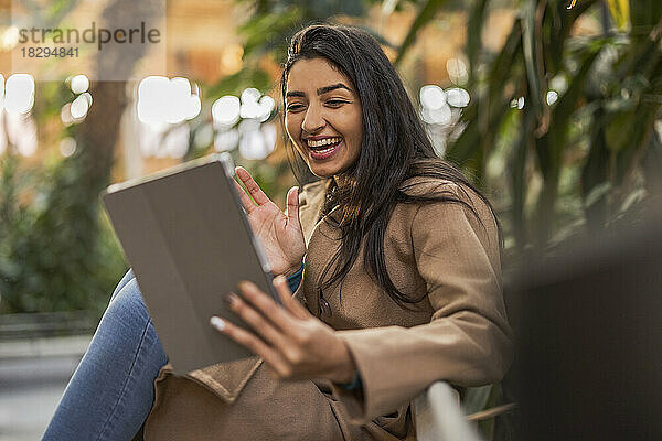 Fröhliche junge Frau winkt bei einem Videoanruf über einen Tablet-PC  der am Geländer sitzt