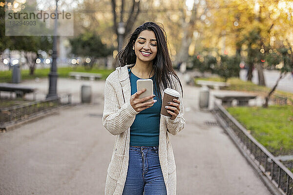 Glückliche Frau mit Einwegbecher und Smartphone im Park