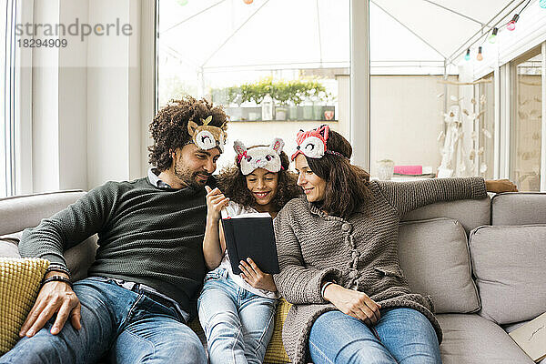 Smiling parents with daughter wearing eye masks and reading book on sofa at home