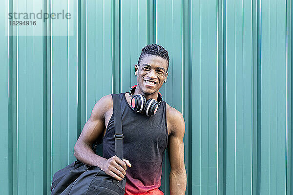 Happy young man with gym bag leaning on green corrugated wall