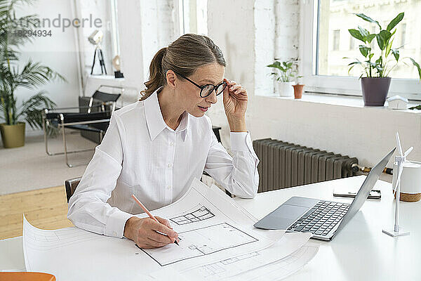 Geschäftsfrau mit Blaupause blickt auf Laptop am Schreibtisch im Büro