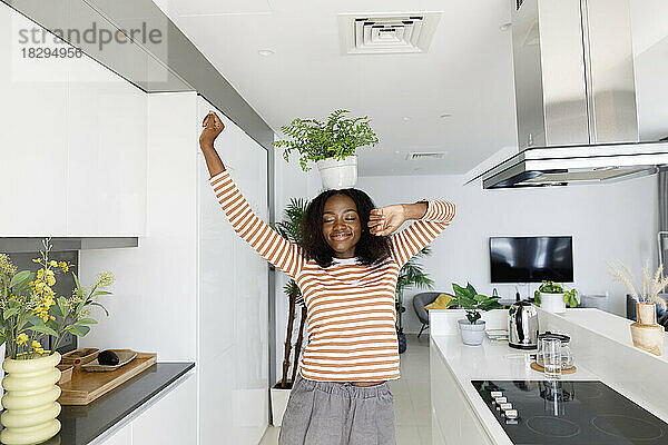 Glückliche Frau mit geschlossenen Augen hält Topfpflanze auf dem Kopf und tanzt in der Küche
