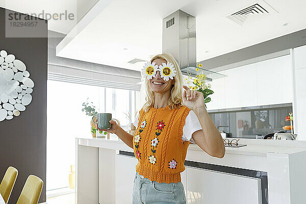 Lächelnde Frau mit Blumensonnenbrille steht mit Teetasse in der heimischen Küche