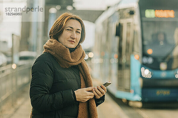 Lächelnde reife Frau steht mit Smartphone an der Straßenbahnhaltestelle