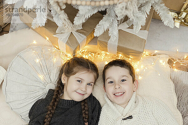 Smiling girl with brother lying by Christmas lights and gifts at home
