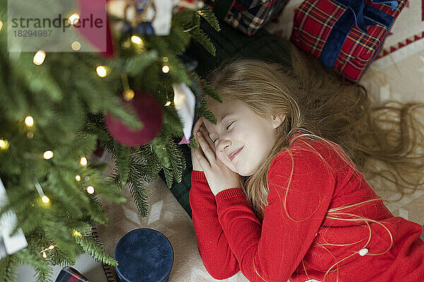 Lächelndes blondes Mädchen entspannt sich zu Hause am Weihnachtsbaum auf dem Teppich