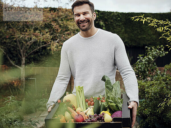 Glücklicher Mann trägt eine Kiste mit frischem Gemüse im Garten
