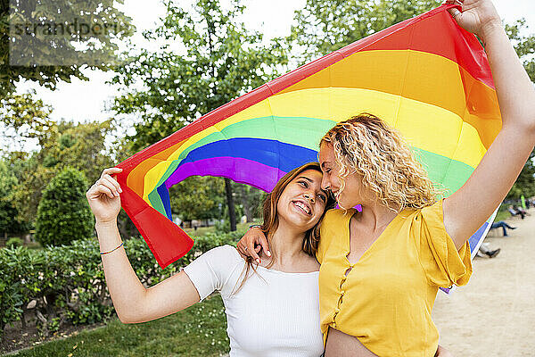 Happy lesbian couple holding rainbow flag embracing each other in park