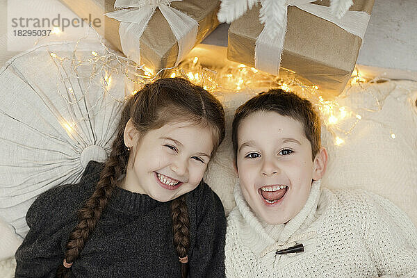 Fröhliches Mädchen und Junge liegen zu Hause bei Weihnachtsbeleuchtung und Geschenken