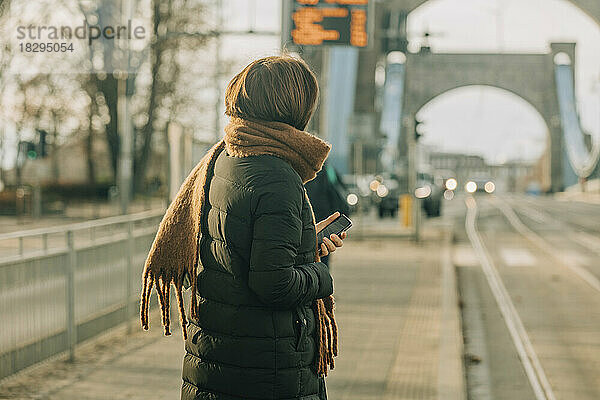 Reife Frau steht mit Smartphone an der Straßenbahnhaltestelle