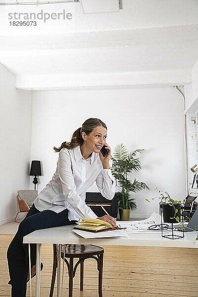 Glückliche reife Geschäftsfrau  die am Schreibtisch im Büro sitzt und über ihr Mobiltelefon spricht