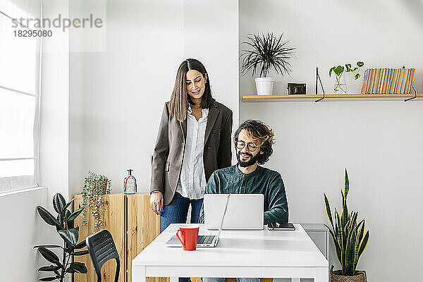 Lächelnder reifer Geschäftsmann teilt Laptop mit Kollegen am Schreibtisch am Arbeitsplatz
