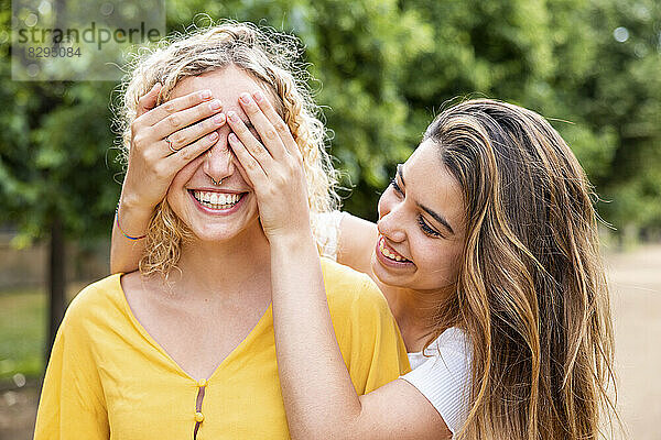 Lächelnde Frau bedeckt die Augen einer Freundin mit den Händen im Park