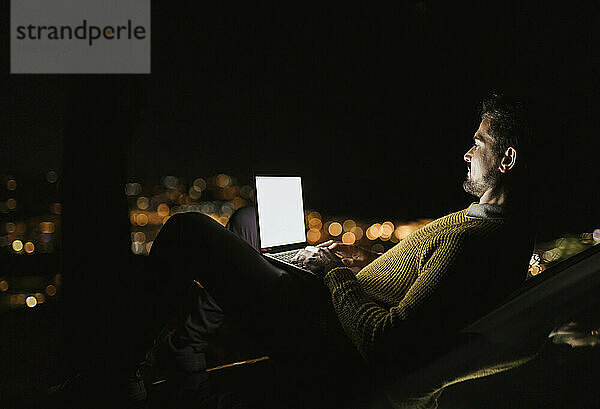 Mann sitzt nachts mit Laptop auf einem Hügel über der beleuchteten Stadt