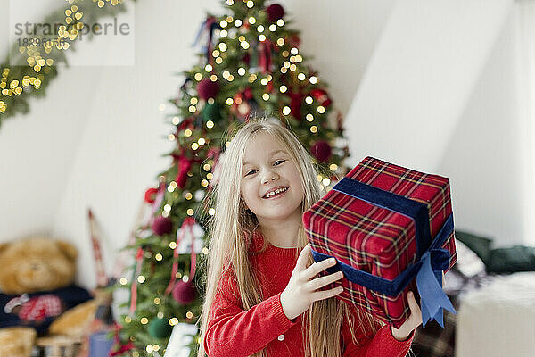 Glückliches blondes Mädchen hält Weihnachtsgeschenk vor dem Weihnachtsbaum