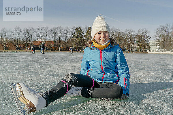 Fröhliches Mädchen mit Strickmütze sitzt auf der Eisbahn