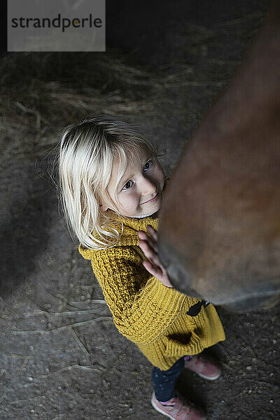 Lächelndes Mädchen streichelt braunes Pferd im Stall