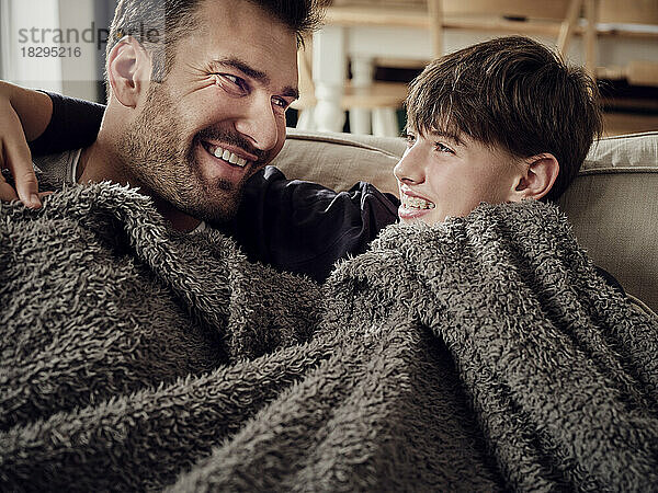 Glücklicher Vater und Sohn kuscheln unter einer Decke auf der Couch im Wohnzimmer