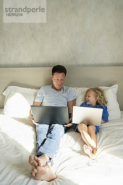 Glückliches Mädchen und Vater mit Laptops auf dem Bett im Schlafzimmer