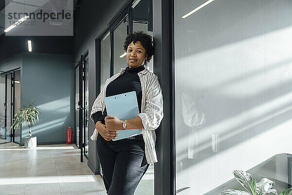 Smiling businesswoman with clipboard leaning on glass in office