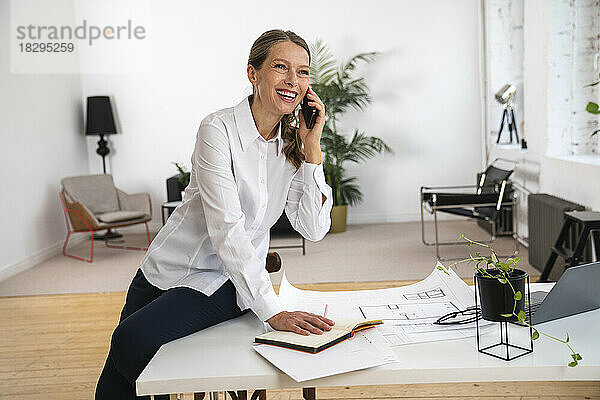 Glückliche reife Geschäftsfrau  die am Tisch im Büro sitzt und über ihr Smartphone spricht