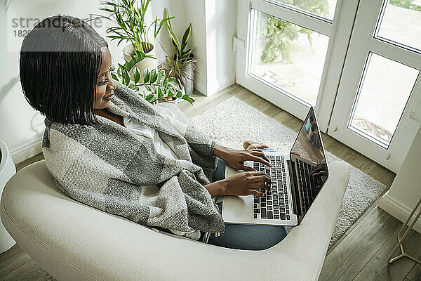 Lächelnde Geschäftsfrau benutzt Laptop im Sessel zu Hause