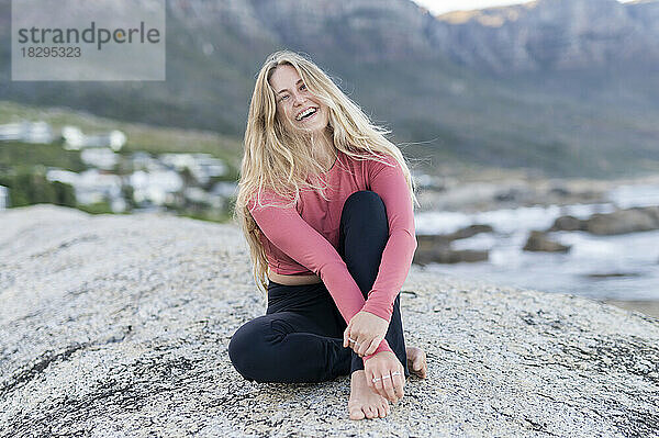 Glückliche Frau mit blonden Haaren sitzt auf einem Felsen am Strand
