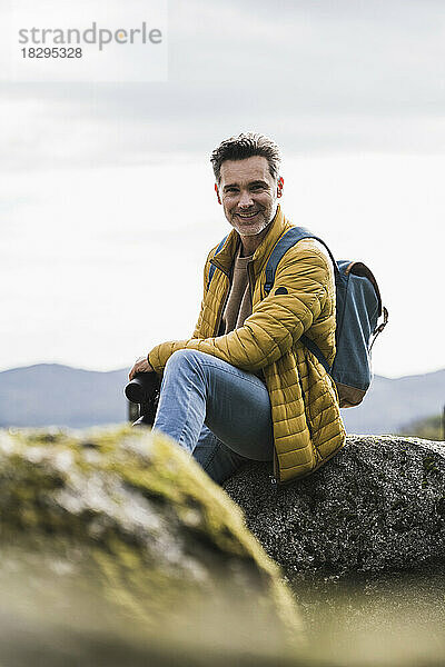 Lächelnder reifer Mann mit Rucksack sitzt auf einem Felsen vor dem Himmel